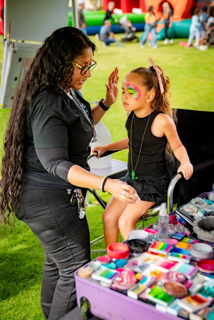 A woman with long dark hair is face painting a young girl. The girl, wearing a black dress and a pink bow, has vibrant colors on her cheeks. They are outdoors on grass, with a table full of paints and brushes nearby.