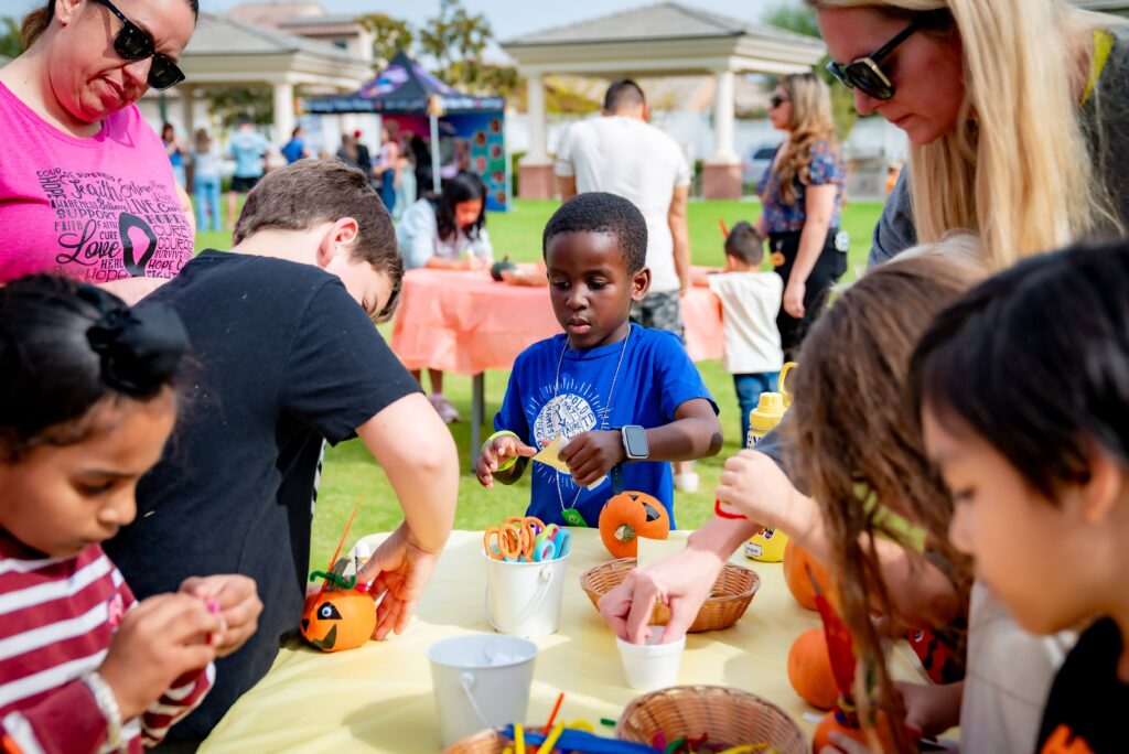 Children gathered around a table outdoors, decorating small pumpkins. They are using markers and craft supplies. Adults are nearby assisting. The scene takes place in a park-like setting with colorful tables and people in the background.