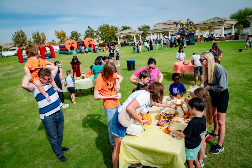 A group of people, including children and adults, gather outdoors at tables to decorate pumpkins. Inflatable pumpkins and a bouncy house are in the background, with trees and picnic shelters under a blue sky. One man carries a child on his shoulders.