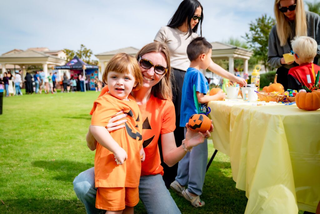 A woman and child in matching orange outfits with jack-o'-lantern designs smile at a festive outdoor event. They hold a painted pumpkin. In the background, adults and children enjoy activities at a table covered with a yellow cloth.