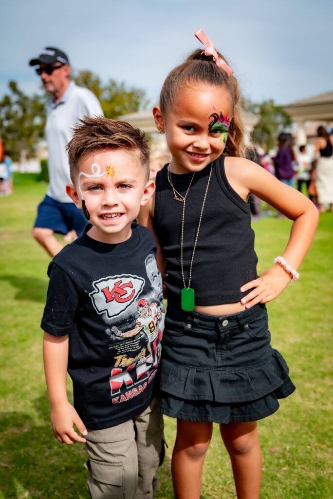 Two children happily pose for a photo at an outdoor event. The boy wears a t-shirt with a sports logo and has face paint of a daisy. The girl, smiling and wearing a black top and skirt, has face paint of a dragonfly. People are in the background.