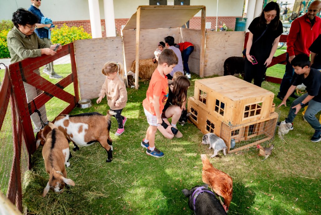 Children and adults interact with various animals, including goats, chickens, and rabbits, in a small wooden enclosure at an outdoor event. Some people are petting the animals, while others observe. A wooden structure is part of the pen.