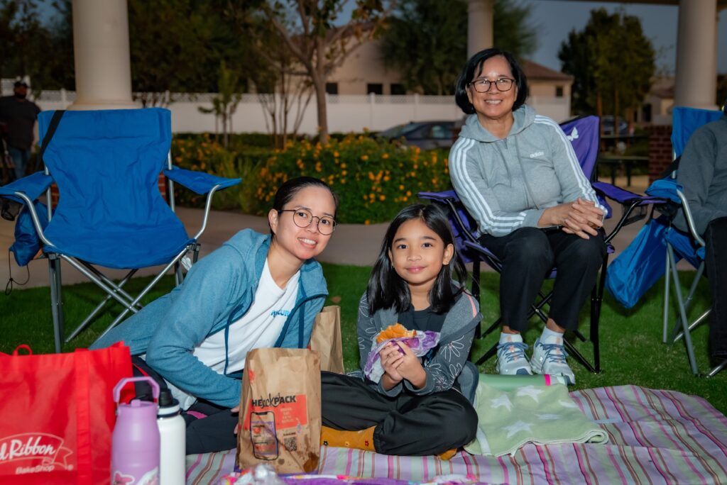 Three people sit together on a picnic blanket in a park. Two women and a young girl smile at the camera. A red bag and a water bottle are on the blanket. The background shows trees and a pavilion.