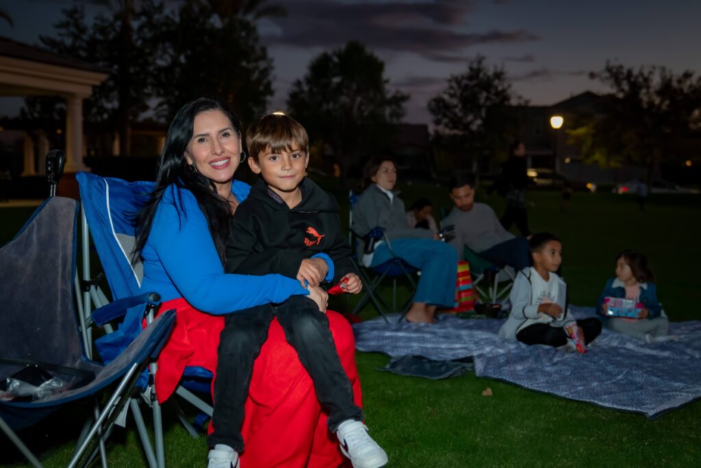 A woman sitting in a camping chair with a boy on her lap. They are outdoors in a park at twilight. Other people are sitting on blankets nearby, some are eating, under a darkening sky.