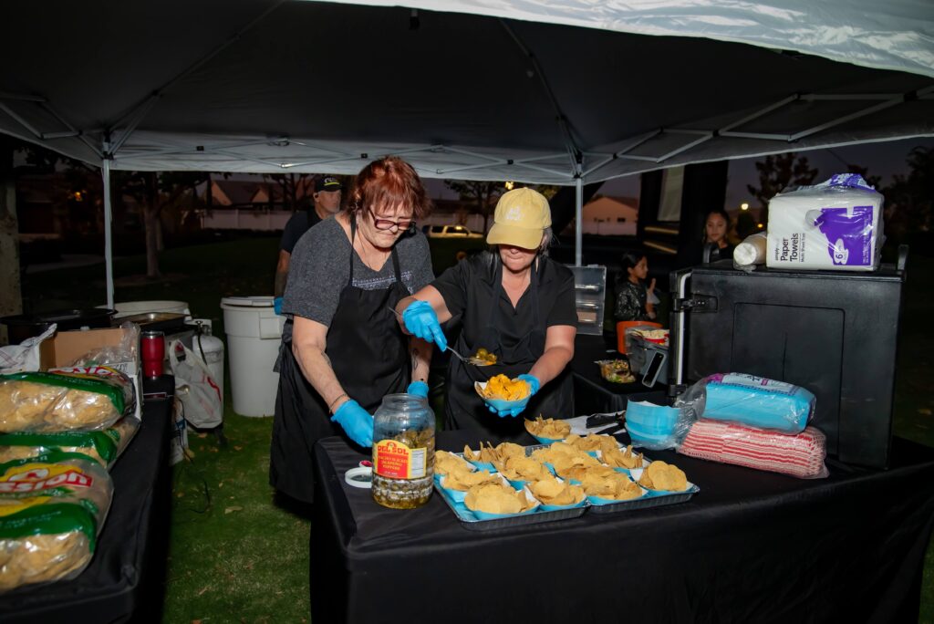 Two people in aprons and gloves prepare food at an outdoor setup under a tent. One holds a tray of nachos, adding toppings, while surrounded by ingredients and supplies. The scene is illuminated against a dusk background.