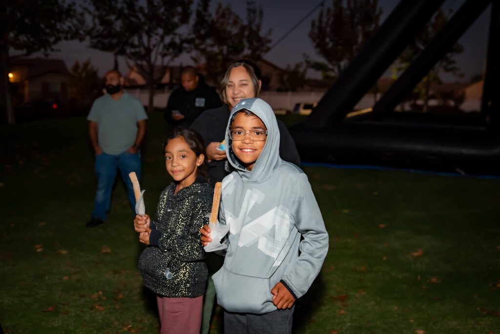 Two children in hoodies smile and hold churros at an outdoor event during the evening. An adult smiles behind them, and a few people stand in the background under a darkening sky.