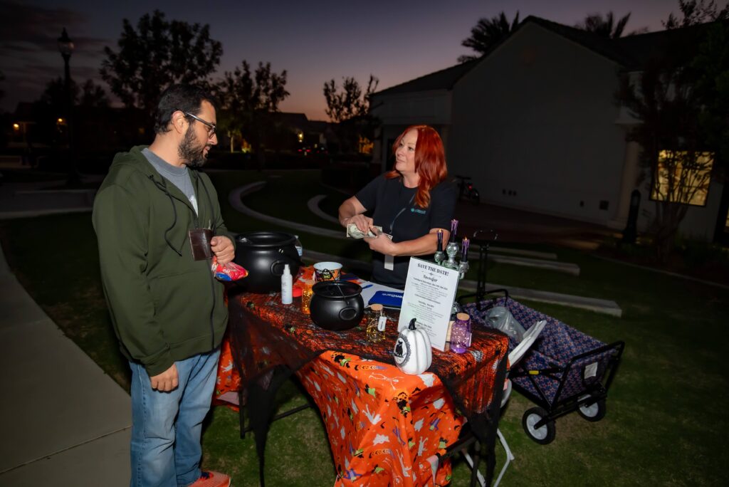 A man and a woman converse at a small outdoor table decorated with Halloween-themed cloth and items, including a cauldron and a sign. The scene is lit by evening light, and a wagon is visible nearby.