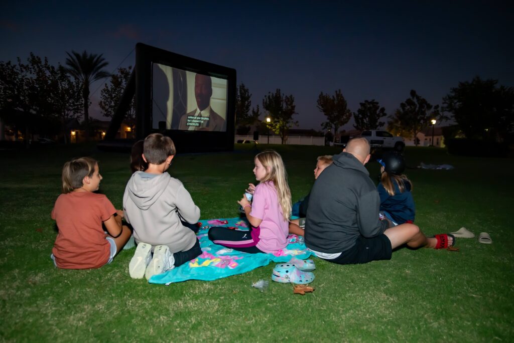 A group of children and a man sit on a blanket on a grassy lawn, watching an outdoor movie on a large inflatable screen at night. Trees and distant lights are visible in the background.