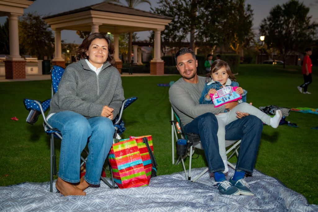 A family sits outdoors on a blanket. A woman in a gray sweater and jeans sits on the left, a man in a gray sweater holds a child with colorful snacks on the right. Behind them are trees and a pavilion in the evening light.