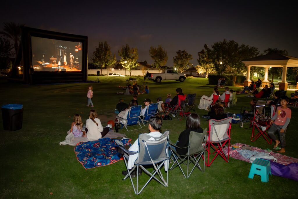 A group of people sit on lawn chairs and blankets watching a movie on a large outdoor screen in a park at night. Trees and a gazebo are visible in the background, and a few children stand nearby.