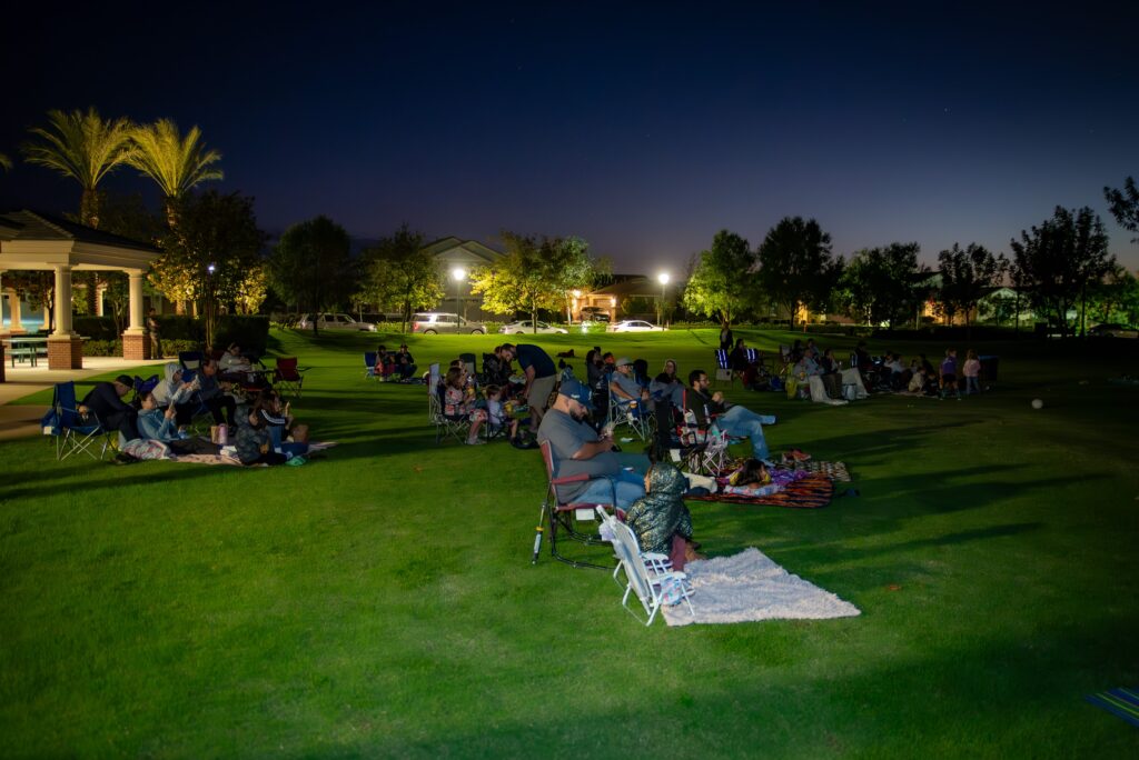 A group of people sits on a grassy field at night, watching an event. Some are seated on chairs while others relax on blankets. The area is dimly lit by surrounding streetlights and trees outline the background.