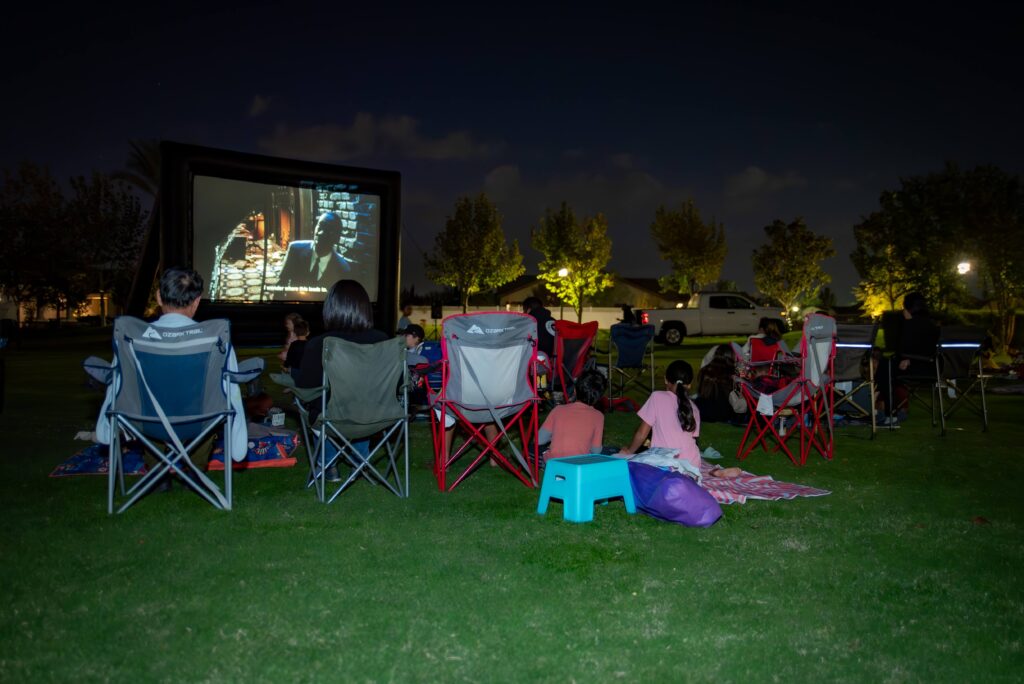 A group of people sitting on chairs and blankets in a park, watching a movie projected on a large outdoor screen at night. The scene is illuminated by ambient light from nearby street lamps and trees.