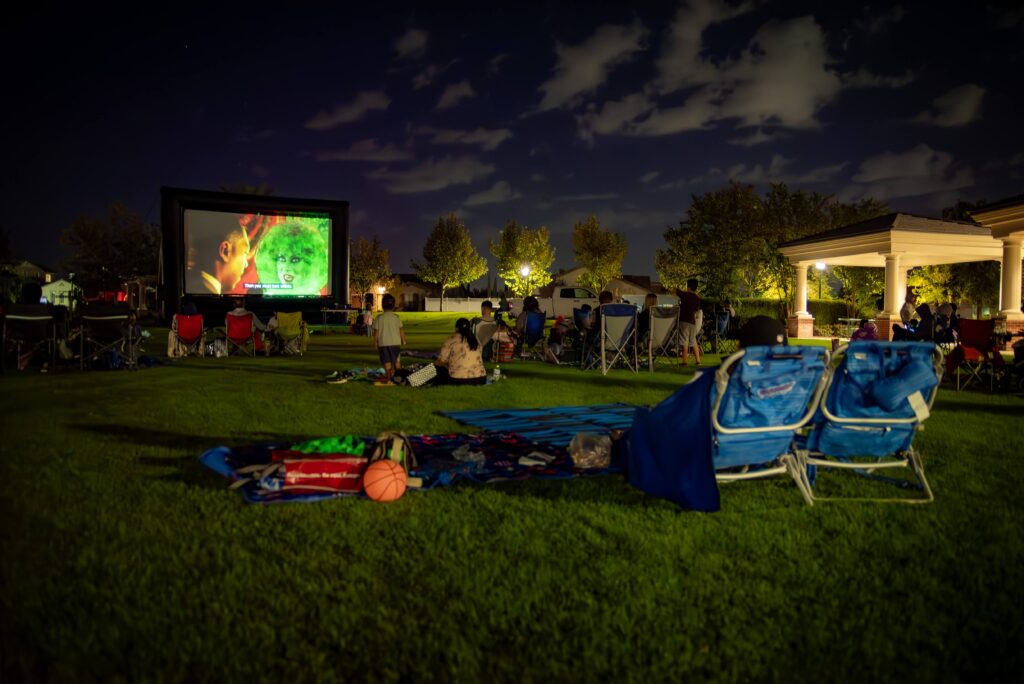 Outdoor movie night scene on a grassy lawn, featuring several people seated in folding chairs watching an illuminated large screen. A basketball and blankets are visible in the foreground under a night sky with scattered clouds.