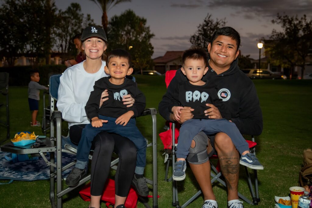 A family of four sits outdoors on lawn chairs during the evening. The parents smile as they hold their two young children, who are wearing black shirts with Halloween-themed designs. Trees and a cloudy sky are in the background.