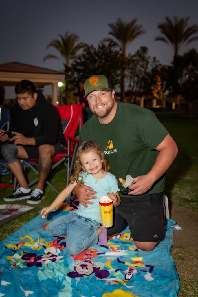 A smiling man kneels beside a young girl on a colorful blanket during an evening outdoor picnic. The girl is holding a popcorn cup, and palm trees are visible in the background. Another person sits nearby.