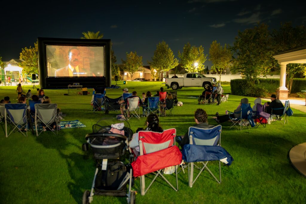 People watching an outdoor movie at night in a park. They are seated in chairs and on blankets on the grass, facing a large inflatable screen showing a film. Trees and lights surround the area, and parked cars are visible nearby.