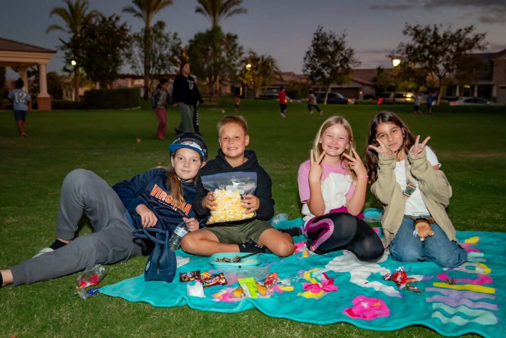 Four children sit on a colorful blanket at an outdoor event, smiling and holding snacks. Two of them make peace signs. It's dusk and people can be seen in the background on a grassy area.