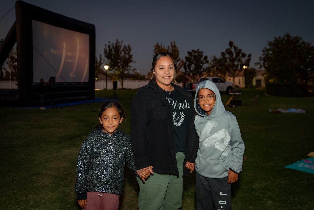 A woman stands with two children on a grassy field at dusk. An inflatable screen in the background shows a movie. The group is dressed in comfortable, casual clothing and smiles softly at the camera. Trees and park lights are visible.