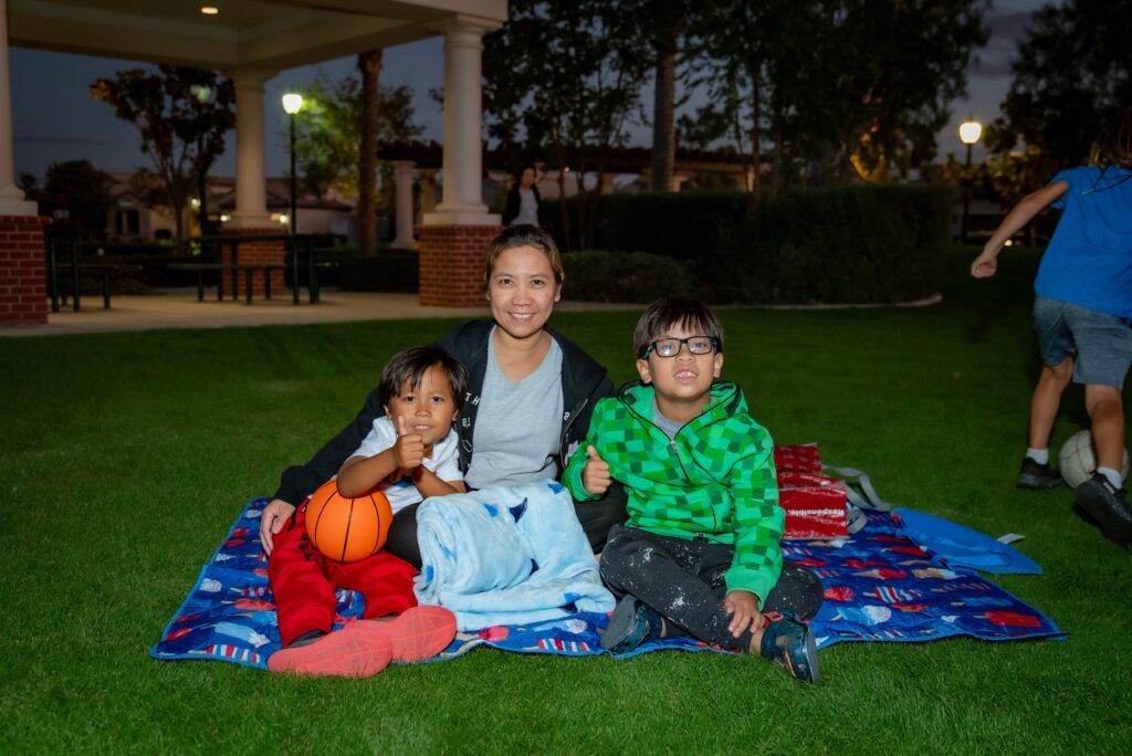 A woman and two children sit on a colorful blanket on the grass during the evening. One child holds an orange basketball. They are smiling at the camera, with a park gazebo and trees in the background. Another child runs nearby.