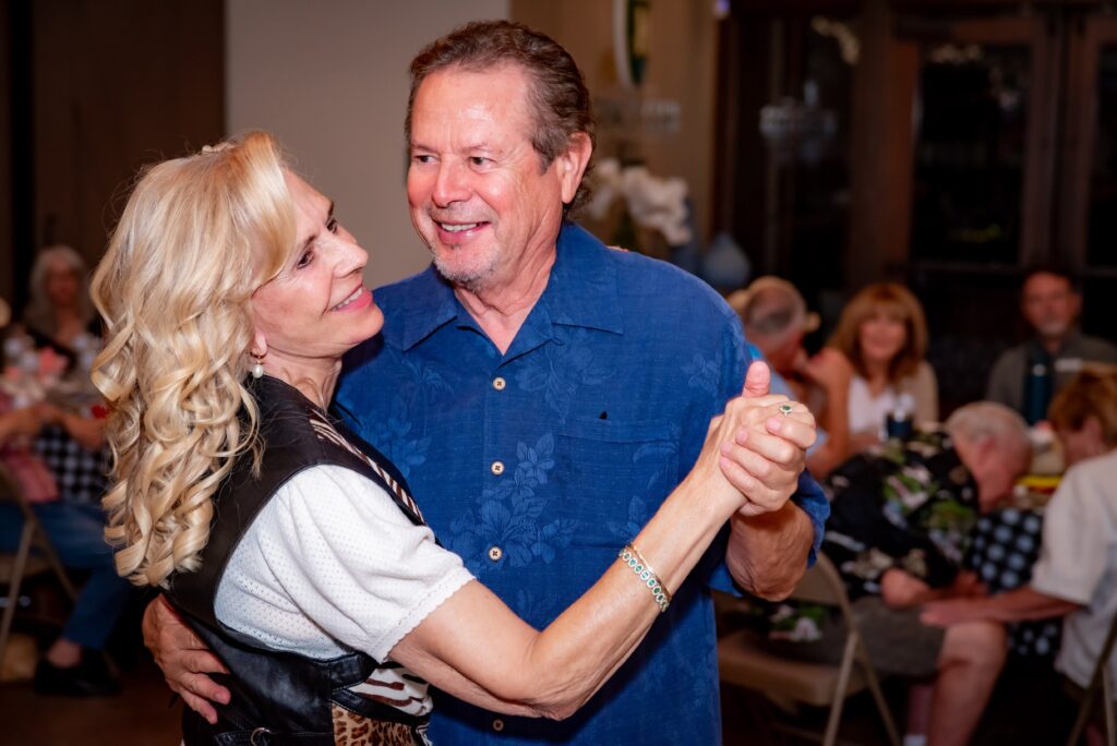 A couple is joyfully dancing together indoors. The woman has long blonde hair and is smiling at the man, who is wearing a blue shirt. They are surrounded by other people sitting at tables in the background.