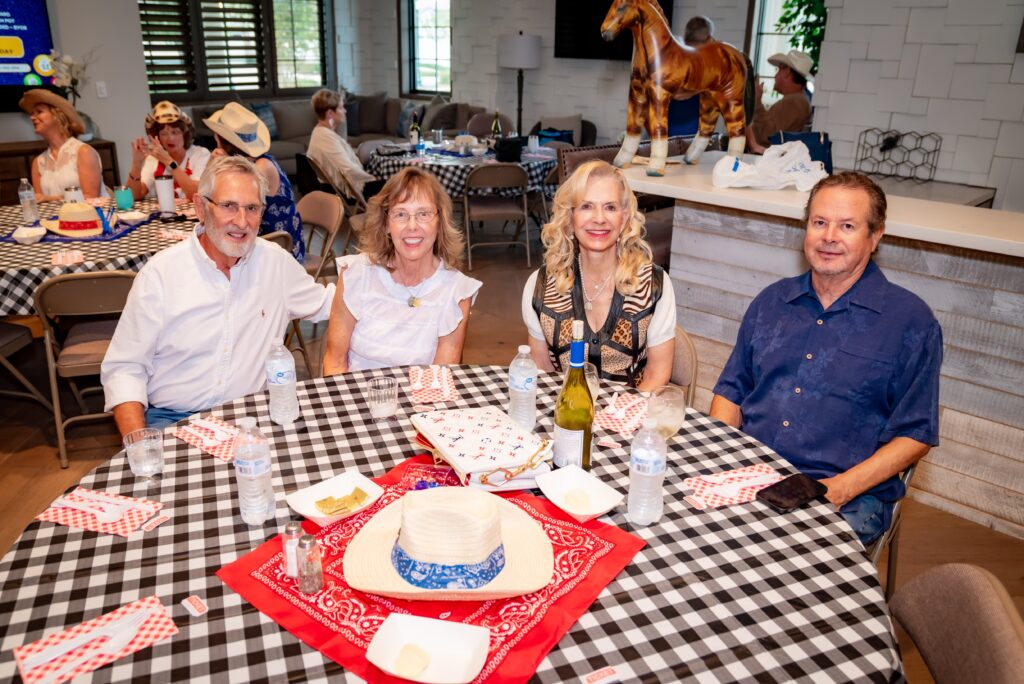 Four people sitting at a table with a checkered tablecloth, wearing casual attire. Water bottles and a wine bottle are on the table. Cowboy hats and bandanas are seen as decorations. A horse statue is in the background. Others are seated or standing nearby.