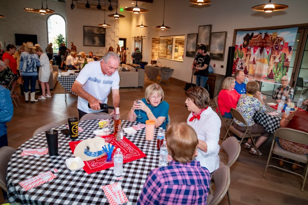 A group of people in a casual indoor gathering, sitting at tables covered with checkered tablecloths. Some are eating and conversing. The room is decorated with western-themed artwork and has a welcoming atmosphere.