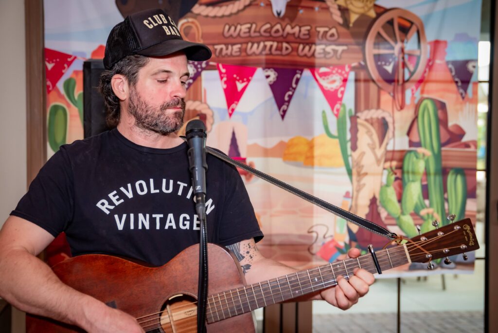 A man wearing a black cap and t-shirt plays an acoustic guitar. In the background, a colorful Wild West-themed decoration with cacti and bandanas is visible.