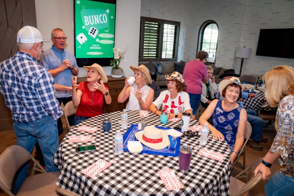 A group of people are gathered around a table with a checkered tablecloth, laughing and talking. Some wear cowboy hats. A poster on the wall reads "Bunco." The atmosphere is lively and social.