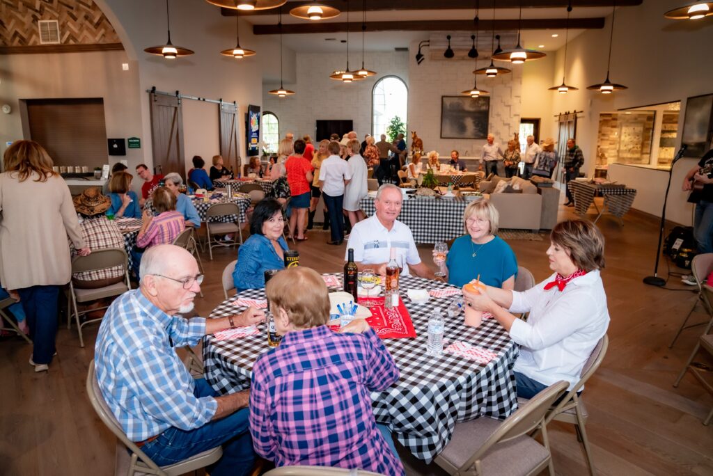 A group of people is gathered in a large, brightly lit room with round tables covered in black and white checkered tablecloths. Some guests are seated and eating, while others are mingling in the background. The atmosphere is social and lively.