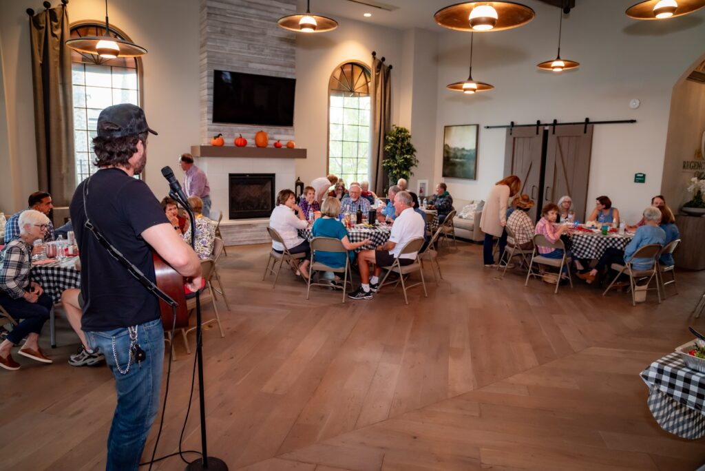 A musician wearing a black shirt and cap sings and plays guitar in a room with tables covered in checkered tablecloths. Several people sit and listen, enjoying a meal. The room is decorated with plants, artwork, and a fireplace with pumpkin decorations.