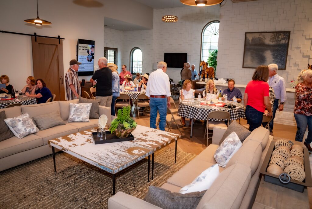 A group of people gather in a spacious living room with sofas and a large coffee table. They are socializing and dining at tables with checkered tablecloths. The room has large windows with arch designs and a large painting on the wall.