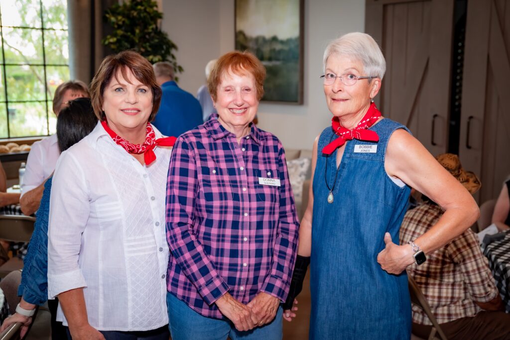 Three older women stand together indoors, wearing casual clothing and red bandanas around their necks. They are smiling, and one has a name badge. The background includes other people and some greenery.