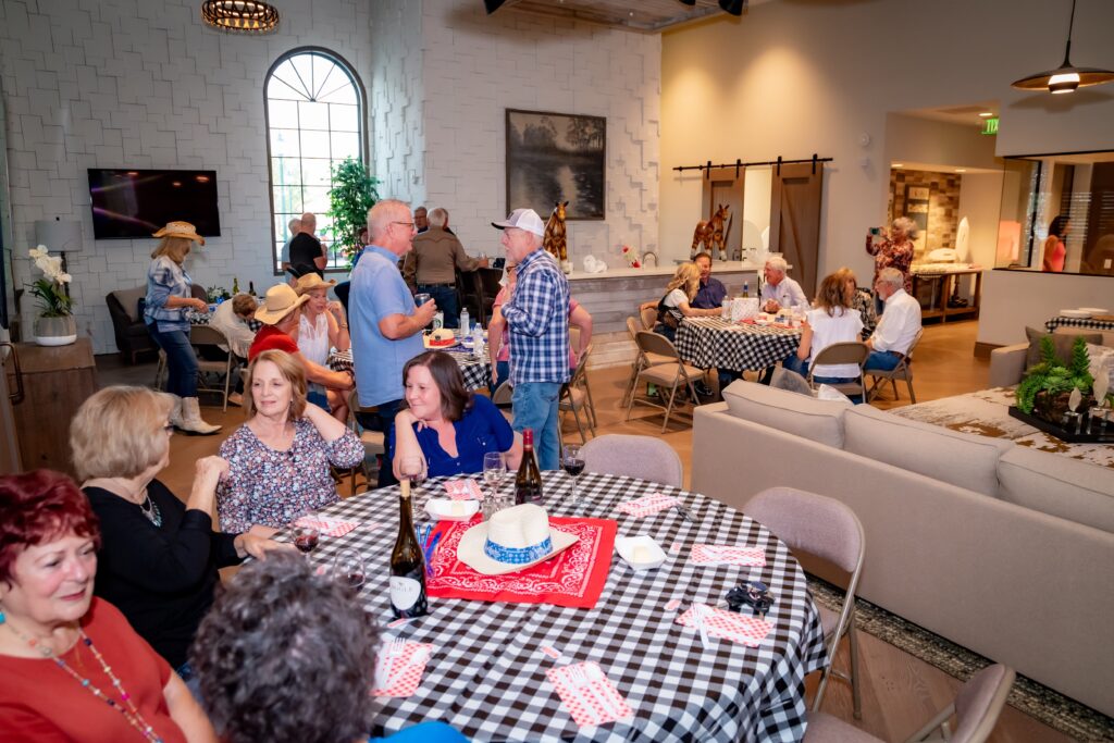 People are gathered in a cozy room with country-style decor. Several groups are seated at tables covered with checkered cloths, chatting and enjoying food. A window with an arched top lets light in, creating a warm atmosphere.