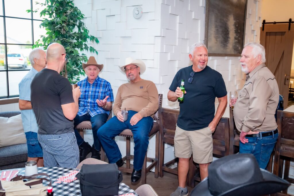 A group of six older men are gathered in a casual indoor setting. Some are sitting on stools, while others stand. They are engaged in conversation, holding drinks, and wearing cowboy hats. A plant and textured white wall are in the background.