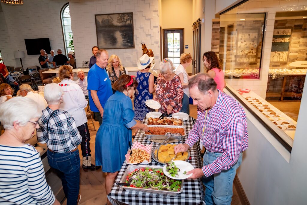 A group of people at a buffet-style gathering indoors. They are serving themselves food from a table covered in a checkered cloth, featuring a variety of dishes. The room is well-lit, and attendees are dressed casually.