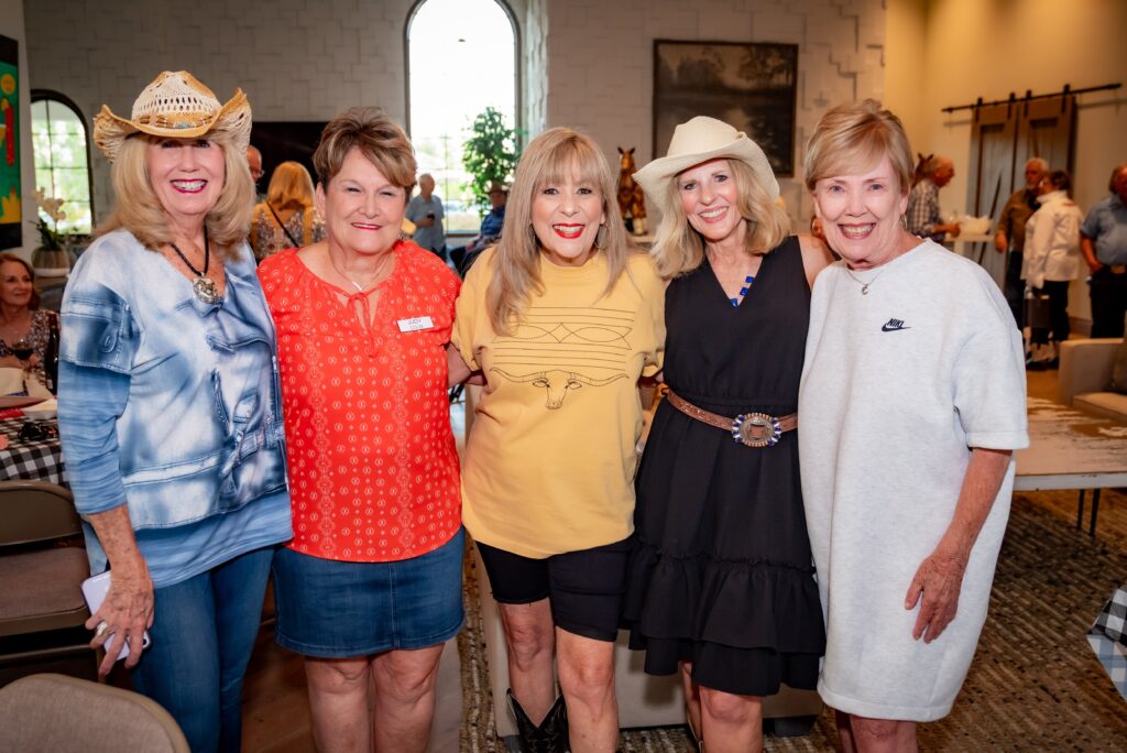 Five women are standing together indoors, smiling at the camera. They are wearing casual and stylish clothing, including cowboy hats and dresses. The background shows a light-filled room with people seated at tables.