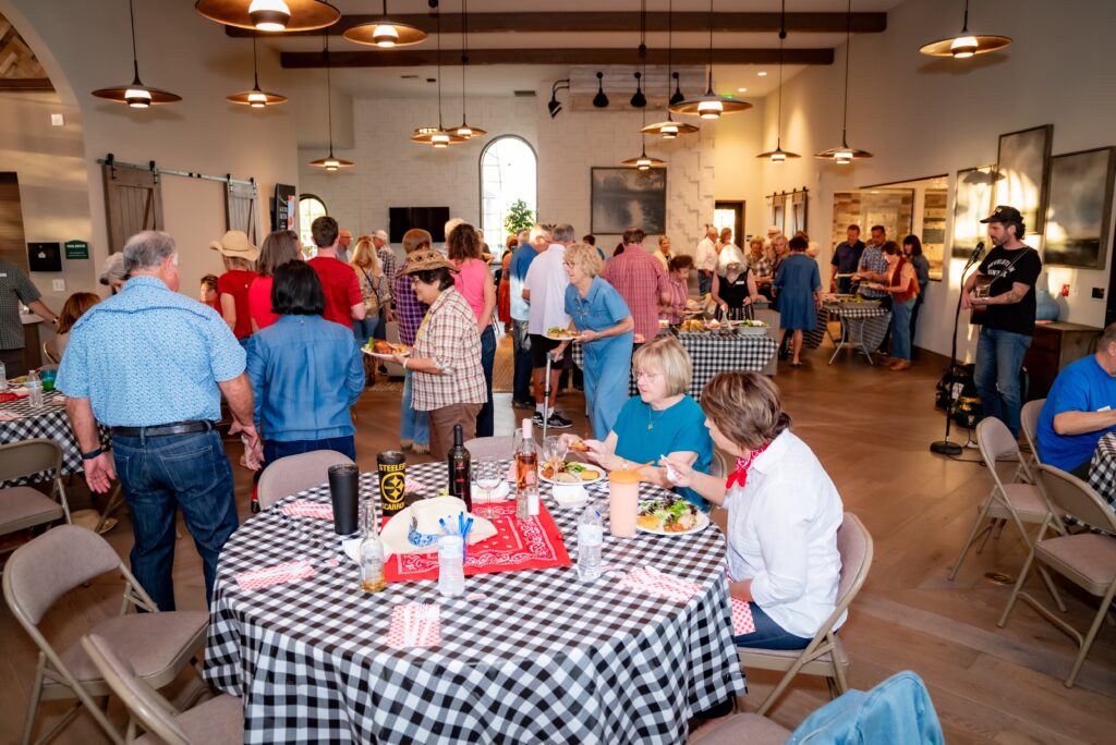 A group of people enjoying a meal in a large room with checkered tablecloths. Some are seated, others are at buffet tables. The room has wooden floors, hanging lights, and artwork on the walls.