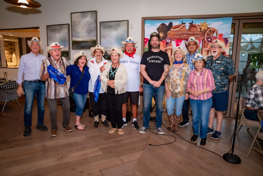 A group of people wearing cowboy hats and Western-themed attire stand together indoors. They are smiling in front of a backdrop with Western motifs. The floor is wooden, and there are framed artworks on the wall.