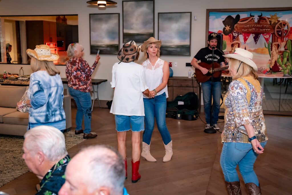 People in cowboy hats and casual attire are dancing indoors. A musician plays guitar in the background. The setting has Western decor with various paintings and props, creating a lively and festive atmosphere.