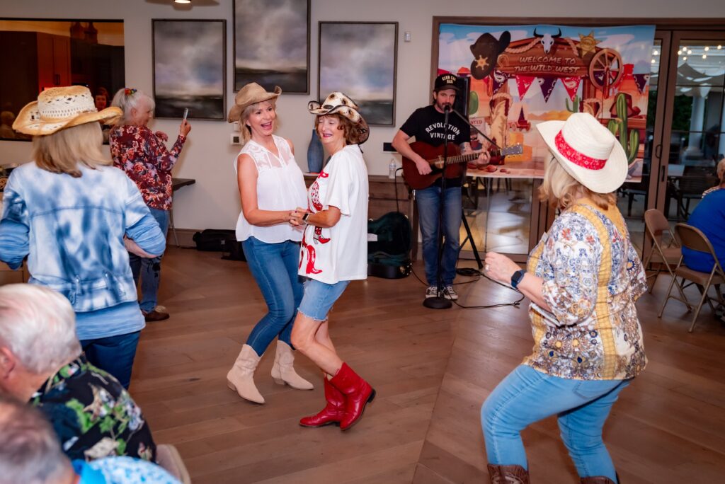 People in cowboy hats are dancing and enjoying music performed by a guitarist in a casual indoor setting. The room is decorated with a Western theme, including a banner and wagon wheel. Everyone appears to be having a good time.