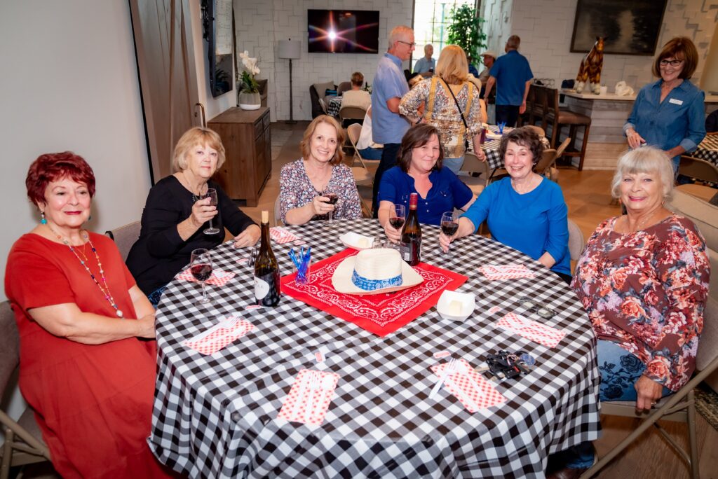 A group of six women seated around a table with a checkered tablecloth, enjoying drinks. The table has wine glasses and a straw hat. In the background, several people stand and converse in a warmly lit room.