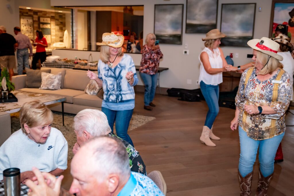 A group of people are indoors at a casual gathering. Some are dancing, wearing cowboy hats and boots, while others are sitting and chatting. The room has a relaxed atmosphere with sofas and artwork on the walls.