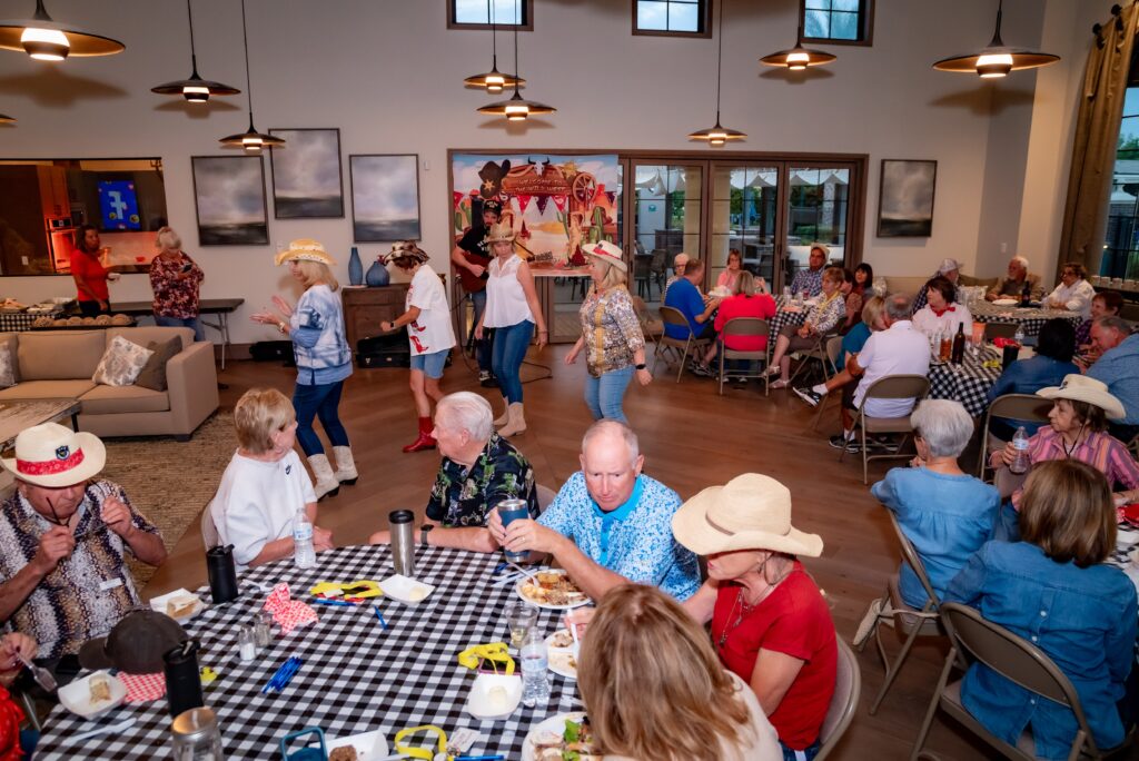 A lively indoor gathering with people seated at tables covered in checkered tablecloths. Attendees, some wearing cowboy hats, are enjoying a meal and conversation, while a few stand dancing. The room is warmly lit and decorated with western-themed elements.