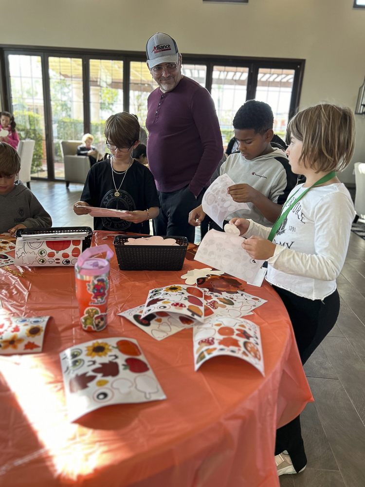 Children and an adult are gathered around a table with a bright orange tablecloth, engaged in a crafts activity involving stickers and paper. The room is well-lit, with large windows in the background letting in natural light.