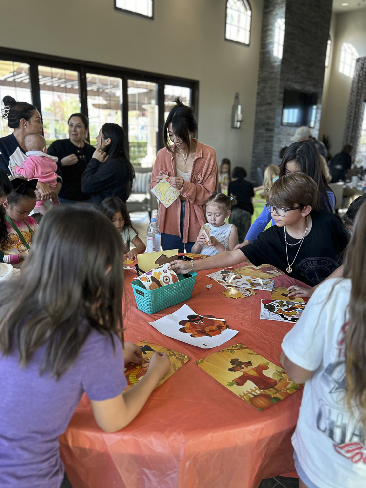 A group of people, including children and adults, are gathered around a table covered with an orange tablecloth. They are engaged in arts and crafts activities, with cut-out figures and papers spread across the table in a room with large windows.