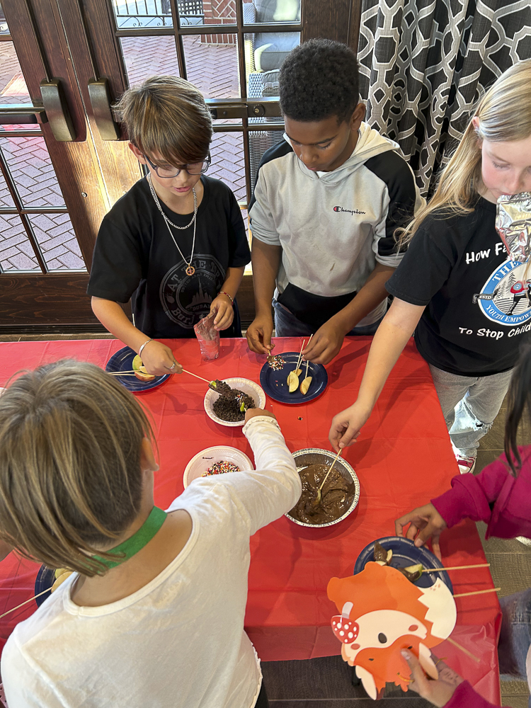 Children are gathered around a table with various toppings, decorating snacks on skewers. The table is covered with a red cloth. Different toppings like chocolate and sprinkles are in bowls. Some kids wear T-shirts with playful designs.