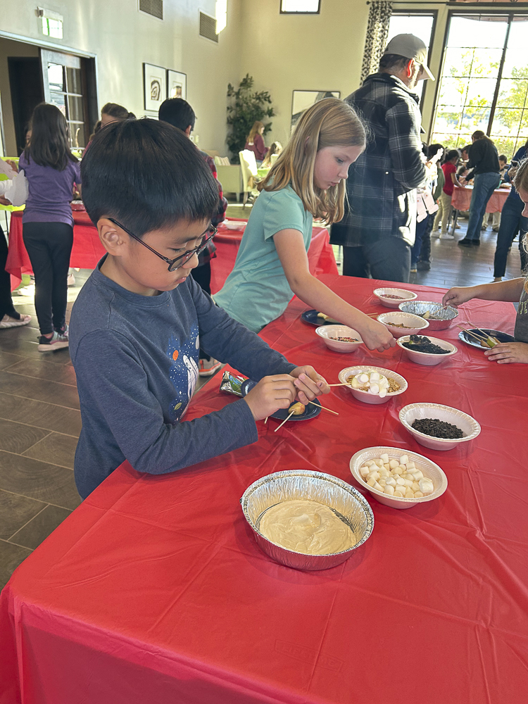 Children are decorating cookies at a table with various bowls of toppings, including marshmallows and chocolate chips. The setting appears to be a community center with other people in the background.