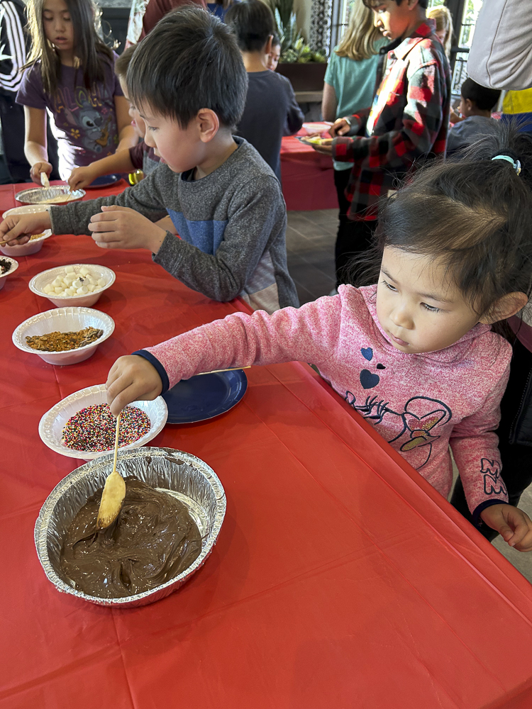 Children are gathered around a red table, decorating dishes with various toppings. A young girl in a pink sweater reaches for a bowl of chocolate spread with a wooden spoon. Other colorful toppings are in bowls nearby.