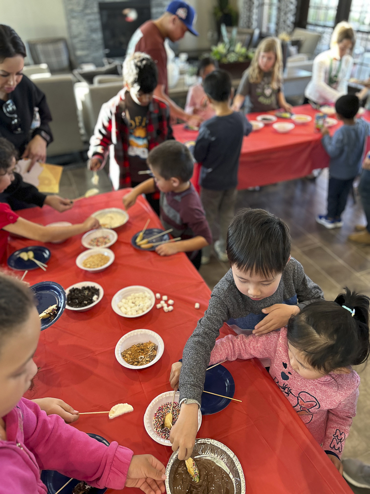 Children gathered around a table with red tablecloths, dipping food items into melted chocolate and adding toppings like sprinkles and marshmallows. Adults are assisting and observing. The scene is lively and interactive.