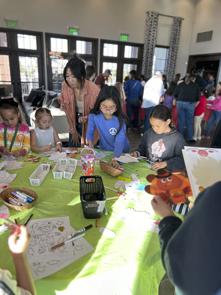 Children and a woman engaged in arts and crafts at a table covered with markers, paper, and supplies. The room is filled with people, and large windows let in natural light.